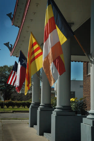 Five poles display different flags outside a building with large columns. The visible flags include U.S., Texas, Vietnam (yellow with red stripes), and a multicolored stripe flag, reminiscent of a Vessak Celebration at a Buddhist Temple. The sky is clear, and the building has architectural details along the roofline.