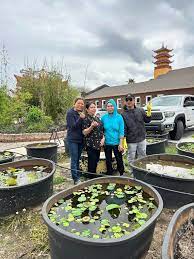 Four individuals stand together, smiling, next to large circular water containers filled with lily pads in an outdoor garden area. There is greenery and a red-roofed building with a pagoda tower in the background. A white truck is also parked nearby, part of the joyful Vessak Celebration at the Buddhist Temple.